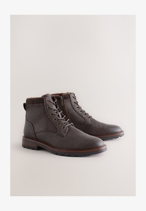 Ruskeat nahkaiset nilkkurit, joissa on teksturoitu kaulus, nauhakiinnitys edessä, sivuvetoketju ja kumipohja, jossa on pitoalueen yksityiskohtia. Varustettu metallisilmukoilla.