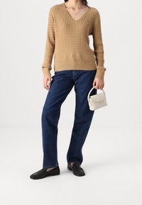 Pull en V beige tricoté, jeans droit bleus, mocassins noirs, et un petit sac à main blanc avec une anse arrondie.