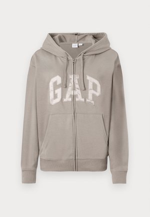 Haut à capuche zippé gris clair avec poches avant, cordon de serrage sur la capuche et grand logo "GAP" sur la poitrine en lettres majuscules.