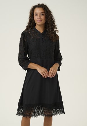 Schwarzes Kleid mit Spitzenoberteil und Dreiviertelärmeln. Verfügt über eine Knopfleiste vorne, einen einfarbigen Rock und Spitzenverzierungen am Saum. Glatte Stofftextur.