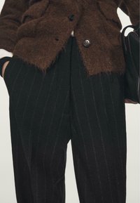 Hnědý chlupatý kardigan přes černé pruhované kalhoty s uvolněným střihem a kapsami. Tkanina má jemný, texturovaný vzhled.