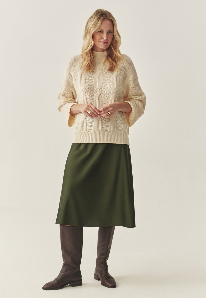 Tekstūra krēmkrāsas kabelīša adījuma džemperis ar brīviem piedurknēm, kombinēts ar zaļu satīna svārku un brūniem ceļgala augstiem zābakiem. Gluda apdare, pieguļošs siluets.