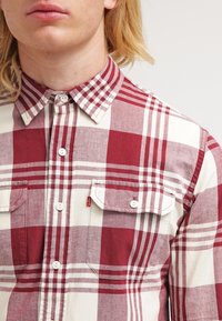 Homme portant une chemise à carreaux rouge et blanche avec deux poches poitrine avant, debout devant un fond clair uni.