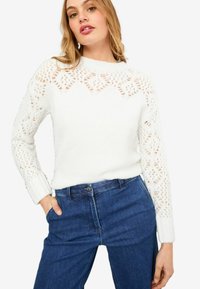 Weißer gestrickter Pullover mit Spitzenmuster an den oberen Ärmeln und am Ausschnitt; kombiniert mit blauen Jeans. Bequeme Textur und taillierte Passform.