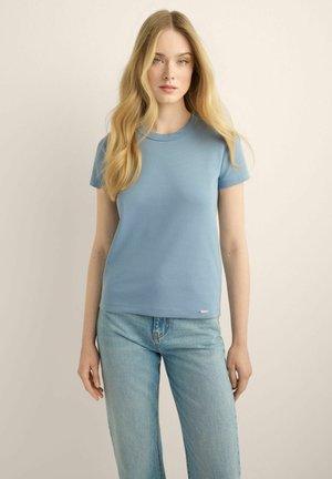 Kobieta z długimi blond włosami, ubrana w jasnoniebieski t-shirt z krótkim rękawem i jasnoniebieskie dżinsy, stojąca na jednolitym tle.