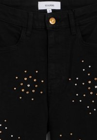 Jeans negros con un botón dorado y adornos dispersos de tachuelas doradas y plateadas en varios tamaños. Presenta bolsillos frontales y una etiqueta.