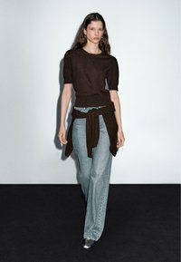 Pull marron à manches courtes avec un accent noué sur un jean large bleu clair et des chaussures argentées métalliques. Le modèle se tient devant un fond uni.