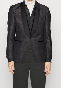 Tuxedo sako ve tmavě šedé látce s jemným leskem, s saténovým šálovým límcem, dvěma předními kapsami a zapínáním na jeden knoflík.