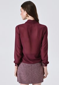 Blusa bordeaux con maniche lunghe, texture morbida e polsini abbottonati, abbinata a una gonna rosa testurizzata con motivo a spina di pesce.