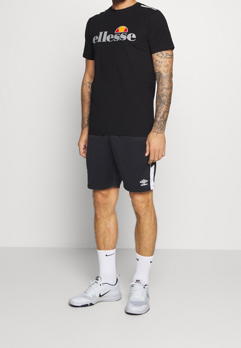 Svart t-shirt med grå "ellesse"-logotyp, kombinerad med grå shorts med vita sidosträck och en vit Umbro-logotyp. Vita Nike-sneakers och strumpor.