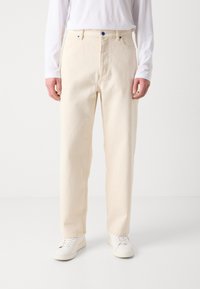 Pantalon beige en coton à jambes droites avec un bouton bleu, poches avant, et texture subtile, associé à des baskets blanches et une chemise à manches longues.