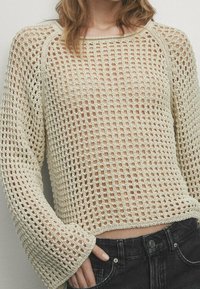 Person, der einen beigen, gehäkelten Pullover mit langen Ärmeln über einem hautfarbenen Top trägt, kombiniert mit hoch taillierten schwarzen Jeans, während die Hand in der Tasche steckt.
