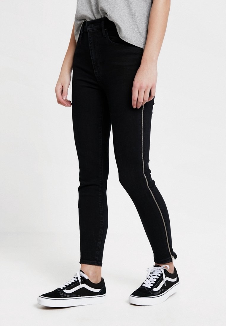 Jean noir taille haute à coupe slim, avec une fermeture éclair latérale et un tissu texturé, associé à des baskets Vans noires.