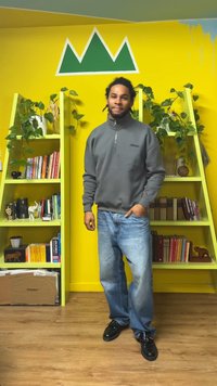 Sudadera gris con cremallera, jeans azules y zapatos negros. De fondo, una pared amarilla con un arte verde en forma de montañas y estantes verdes.