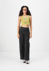 Young woman wearing purple sunglasses, green cropped tank top, black wide-leg pants, and white sneakers, standing against a white background.