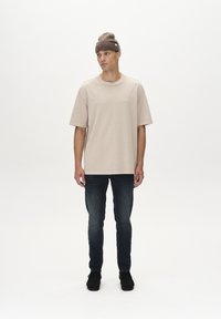 Beige oversize t-krekls ar apaļu kakla izgriezumu, apvienots ar tumši ziliem skinny džinsiem. Modelis valkā gaiši brūnu adītu cepuri un melnas kurpes.