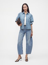 Femme debout face à l'avant, portant une chemise en jean bleu clair et un jean, des sandales à talons noires à brides et tenant un sac marron sous le bras.