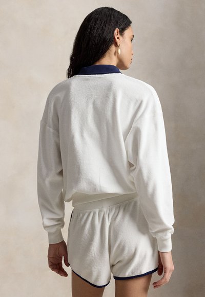 Hosszú sötét hajú nő, fehér hosszú ujjú felsőt és fehér, sötétkék szegélyű rövidnadrágot visel, áll, és a kamerától távolabb néz.