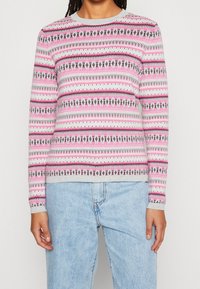 Pull en maille à manches longues, de couleur gris clair, orné de motifs géométriques roses, noirs et blancs. Encolure ronde et poignets côtelés.