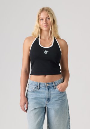 Lyshåret kvinde iført en sort halter-croptop med hvid kant og stjerne-design, parret med lyseblå jeans med høj talje, stående mod grå baggrund.