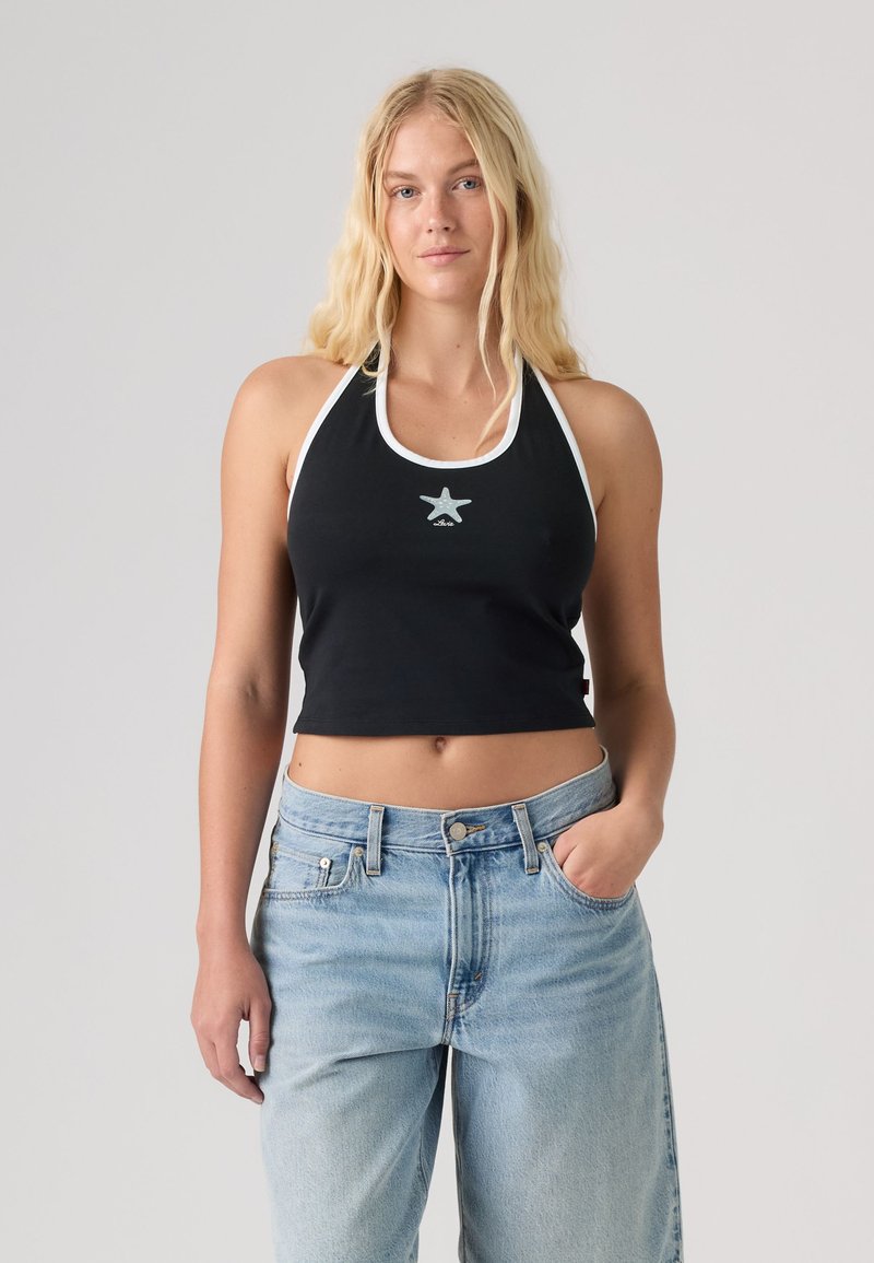 Ξανθιά γυναίκα φοράει μαύρο τοπ halter crop με λευκή επένδυση και σχέδιο αστέρα, σε συνδυασμό με ανοιχτό μπλε ψηλόμεσα τζιν, στέκεται μπροστά σε γκρίζο φόντο.