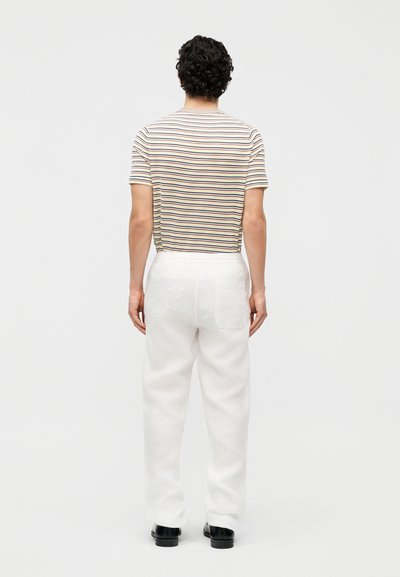 Άνδρας με σγουρά μαλλιά, φοράει μπλούζα με κοντά μανίκια σε μπεζ και μαύρες ρίγες, λευκό παντελόνι και μαύρα παπούτσια, στέκεται γυρισμένος με την πλάτη.