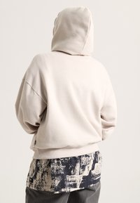Light beige hoodie with a spacious fit, soft texture, and a hood. Underneath, a patterned black and cream layered shirt hem is visible.