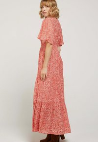 Femme aux cheveux blonds portant une robe maxi à motifs paisley rouge et des bottes brunes, posée debout et regardant par-dessus son épaule.