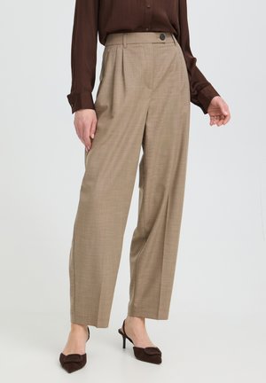 Personne portant un pantalon beige taille haute et à jambes larges, un chemisier marron à manches longues et des talons marron foncé, debout devant un fond uni.