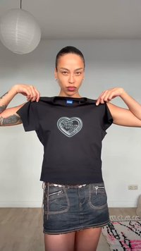 Γυναίκα με μαλλιά ανακατεμένα προς τα πίσω κρατάει μαύρο T-shirt Hugo με λογότυπο καρδιάς, φορώντας κοντή σκουρόχρωμη τζιν φούστα σε έναν μίνιμαλ χώρο.