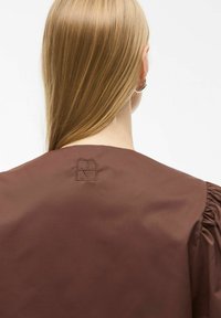 Pruun satiintop, millel on puhv-näppu varrukad ja tagaküljel kaeluses peene logo tikand. Sile tekstuur, minimalistlik disain.