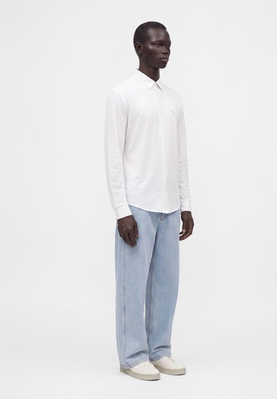 Homme debout portant une chemise blanche à manches longues boutonnée, un jean large bleu clair et des baskets blanches sur un fond uni.