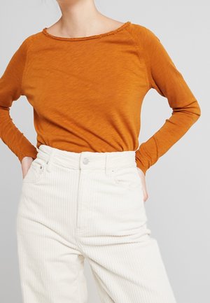 Personne portant une chemise orange brûlé à manches longues rentrée dans un pantalon en velours côtelé blanc taille haute, les mains dans les poches.
