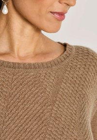 Eine Frau trägt einen braunen strukturierten Strickpullover und einen goldenen Ohrring mit einer hängenden weißen Perle, sichtbar von Hals bis Kinn.
