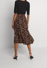 En person står med ryggen mot kameran, iklädd en figursydd svart topp och en leopardmönstrad midikjol, tillsammans med svarta klackar.