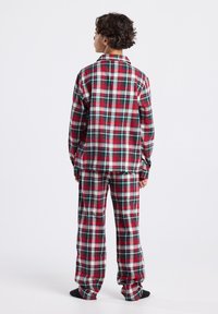 Person standing facing away wearing red, green, and white plaid long-sleeve top and matching loose pants with black socks on white background.