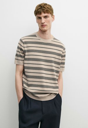 Giovane uomo che indossa un maglione beige a maniche corte con sottili strisce nere orizzontali e pantaloni scuri, in piedi con le mani nelle tasche davanti a uno sfondo bianco.