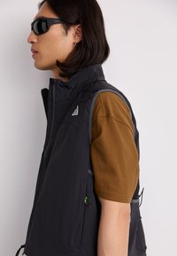 Black sleeveless vest with a high collar, gray accents, and pockets, paired with a brown short-sleeve shirt. Features a logo on the chest.