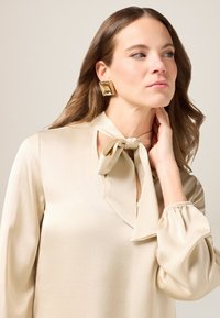 Seidenbluse in hellem Beige mit einem Schleifen-Halsausschnitt, langen Puffärmeln und einer Schlüsselöffnungsdetails. Goldene quadratische Ohrringe setzen einen Kontrast.