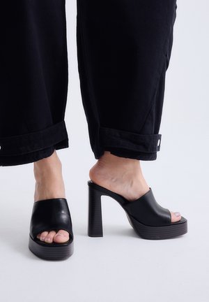 Pies con zapatos de plataforma negros de punta abierta y tacones gruesos de bloque, combinados con pantalones negros con puños sobre un fondo neutro.