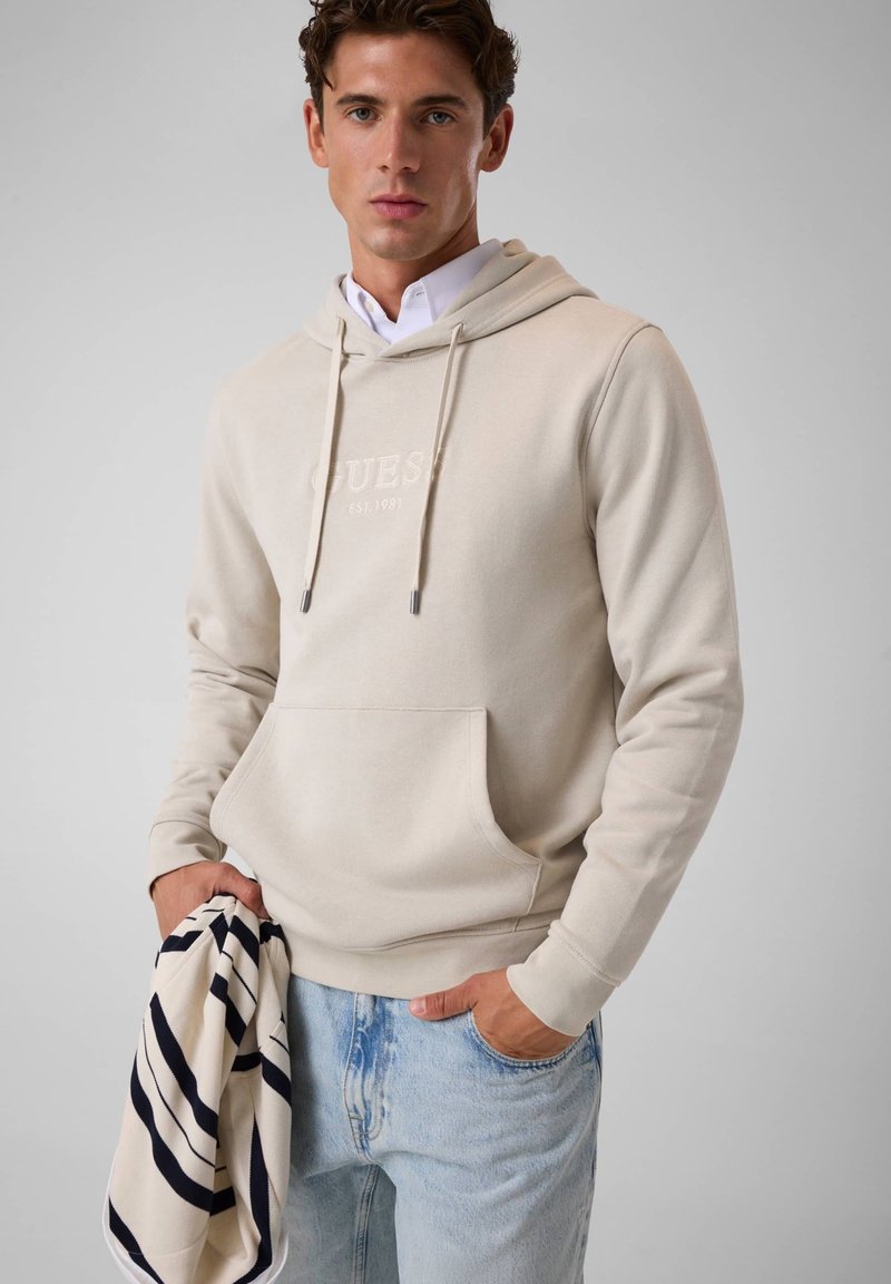 Jeune homme portant un sweat à capuche beige et un jean bleu clair tenant un vêtement rayé noir et blanc devant un fond uni.
