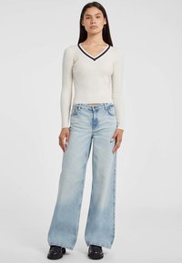 Donna in piedi contro uno sfondo bianco, indossa un maglione crema a costine con scollatura a V e rifiniture blu navy, jeans larghi azzurri e scarpe nere.