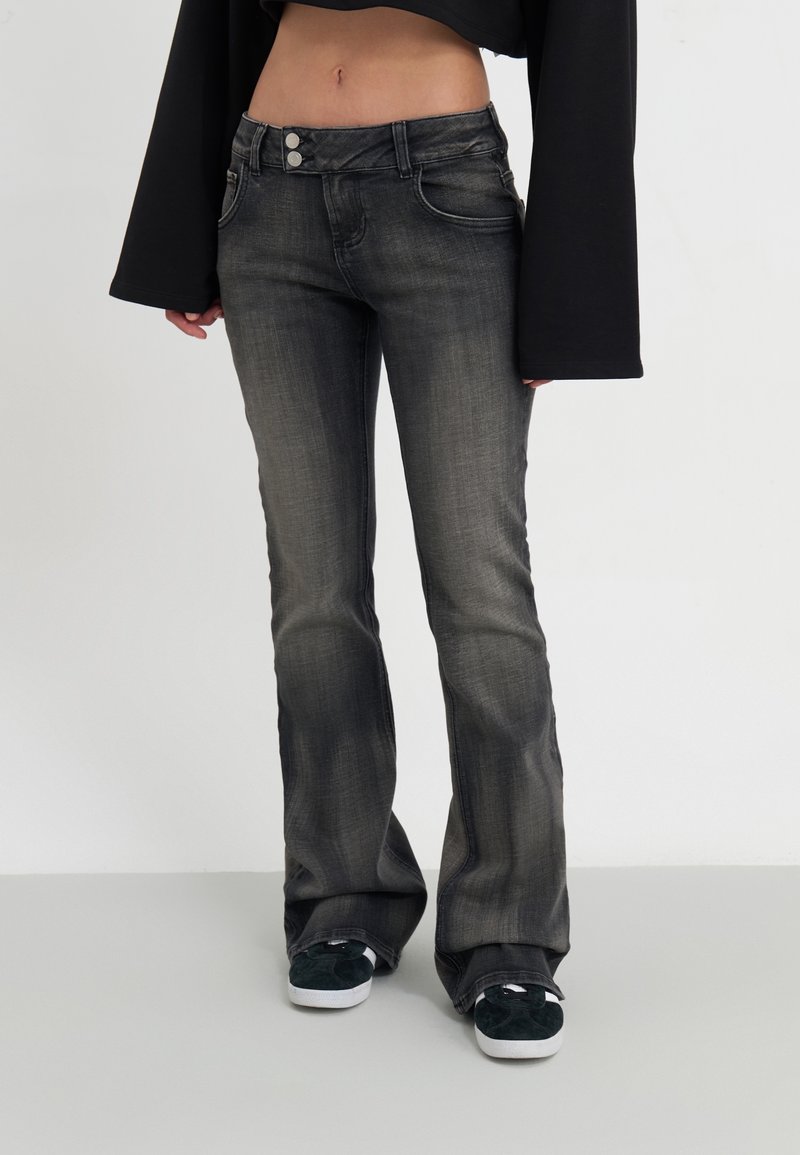 Jeans acampanados en denims gris oscuro con un acabado desgastado, que presentan dos botones frontales y bolsillos delanteros, combinados con zapatillas negras.