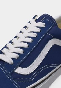 Gros plan d'une basket bleu marine avec des lacets blancs et un détail en forme de vague blanche sur le côté, montrant les coutures et une partie de la semelle blanche.