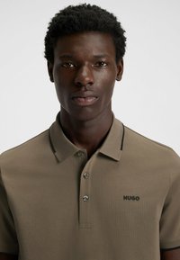 Kortärmad khakifärgad polo med svarta krageinslag, med en treknappars placket och broderad logga på vänster bröst.