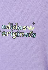 Liliowy bawełniany materiał z nadrukowanym logo "adidas Originals" w czerni i niebieskim, z zielonym akcentem oraz małymi elementami graficznymi.