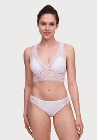 Bralette bianco in pizzo con design a V, elastico e coppe morbide. Slip abbinato in pizzo bianco con vita semi-trasparente e aperture per le gambe.
