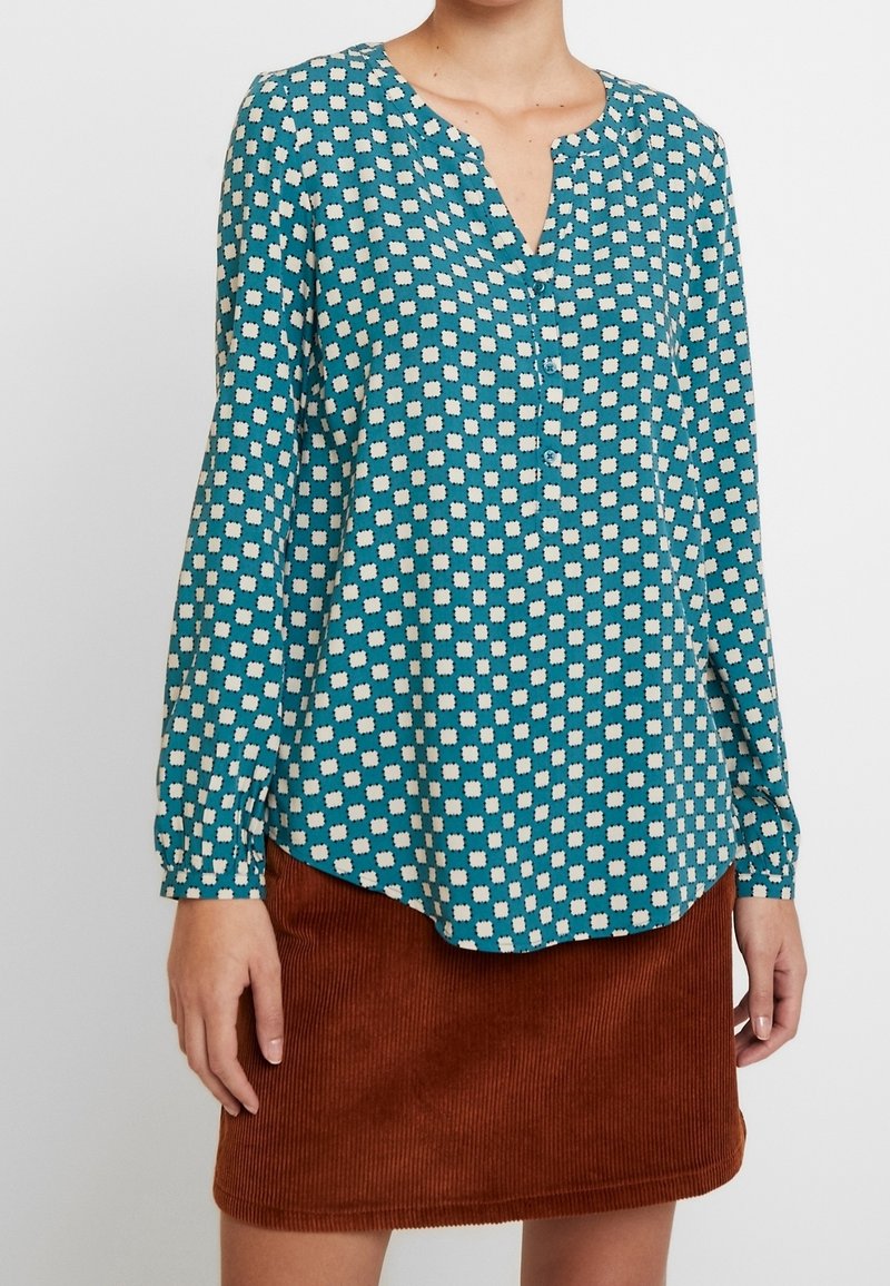 Femme portant un chemisier bleu sarcelle à manches longues avec des motifs carrés blancs et une jupe en velours côtelé marron sur un fond uni.