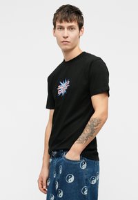 Noor mees kannab musta graafilise t-särki ja siniseid džinsse, poseerides ühe käega taskus ning paistab tema randmetatuu.