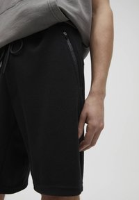 Shorts en mesh noir présentant un tissu texturé avec une taille à cordon de serrage et des poches latérales zippées. Portés avec un haut gris à manches courtes.